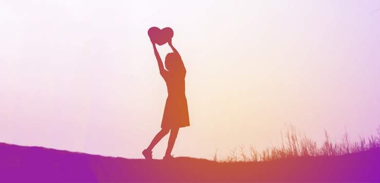 A person in an orange dress holds up a red heart while standing on a hill at sunset.