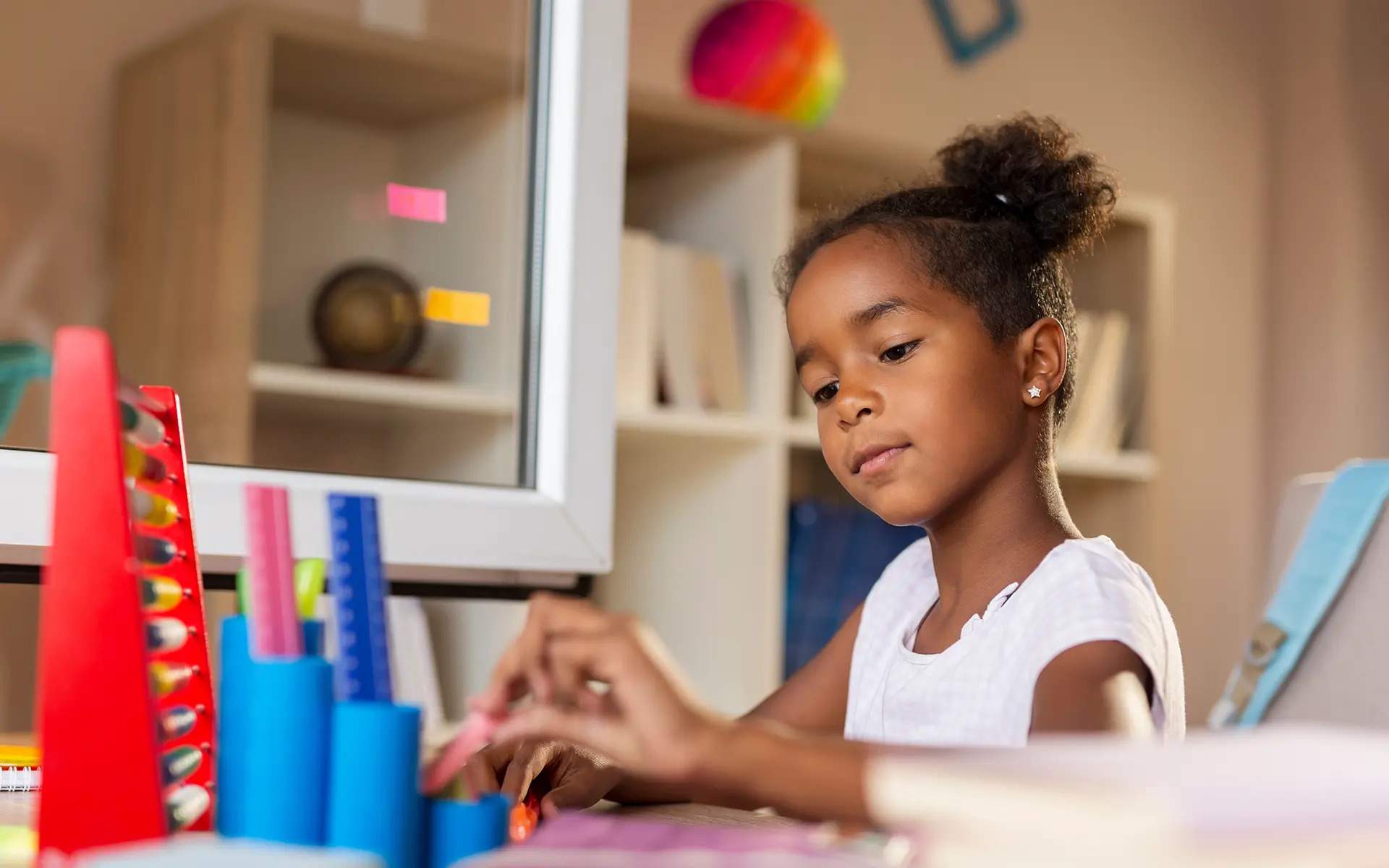A young girl with curly hair tied in a bun is sitting at a desk, engaging with colorful learning materials. She appears focused as she interacts with educational tools like rulers and stacking objects. A bookshelf with books and decorative items is visible in the background.