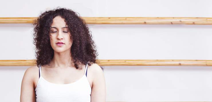 Woman with curly dark hair wearing a white sleeveless top against a white wall with wooden shelving.