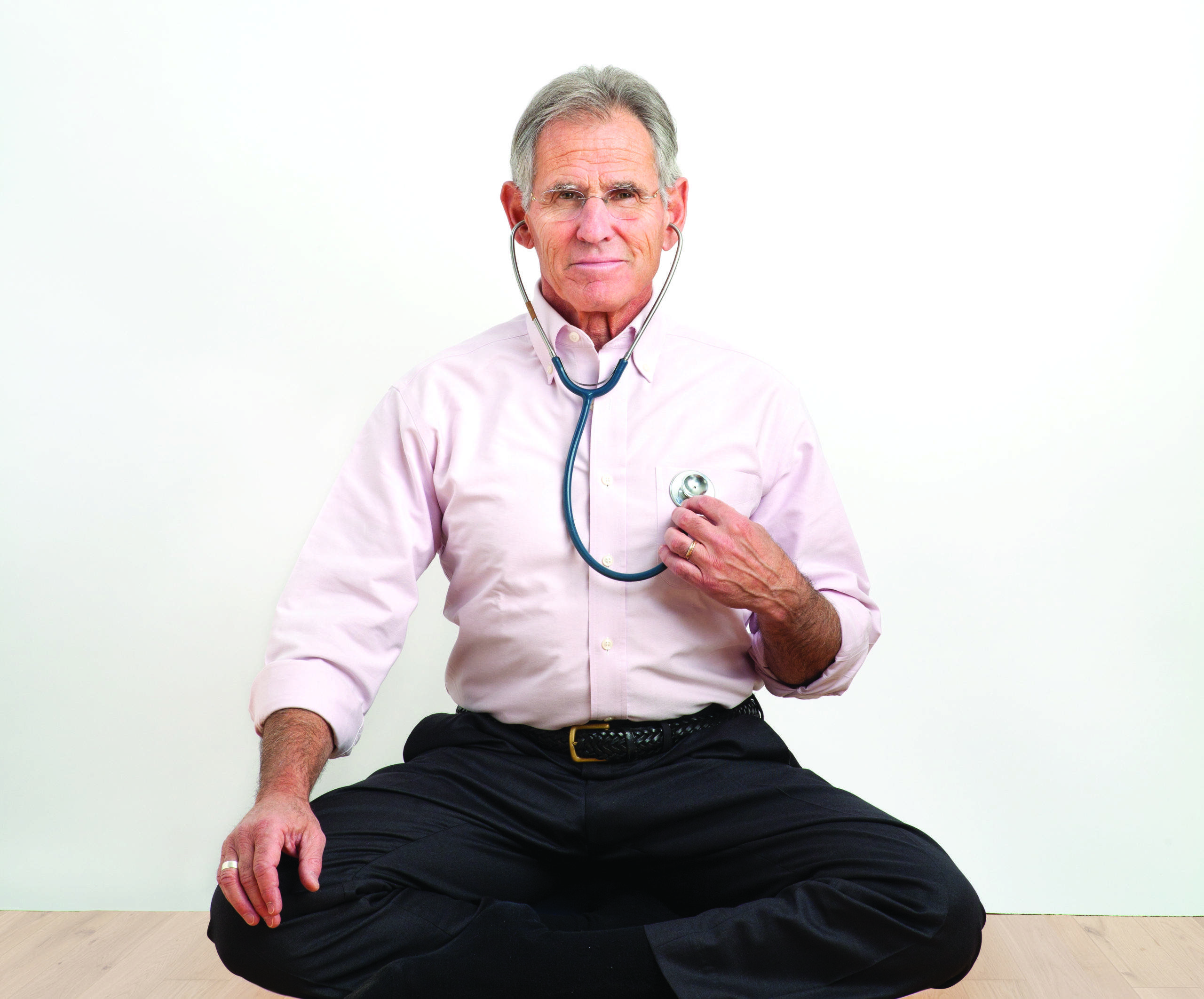 A doctor in a pink shirt holding a stethoscope while sitting against a white background.