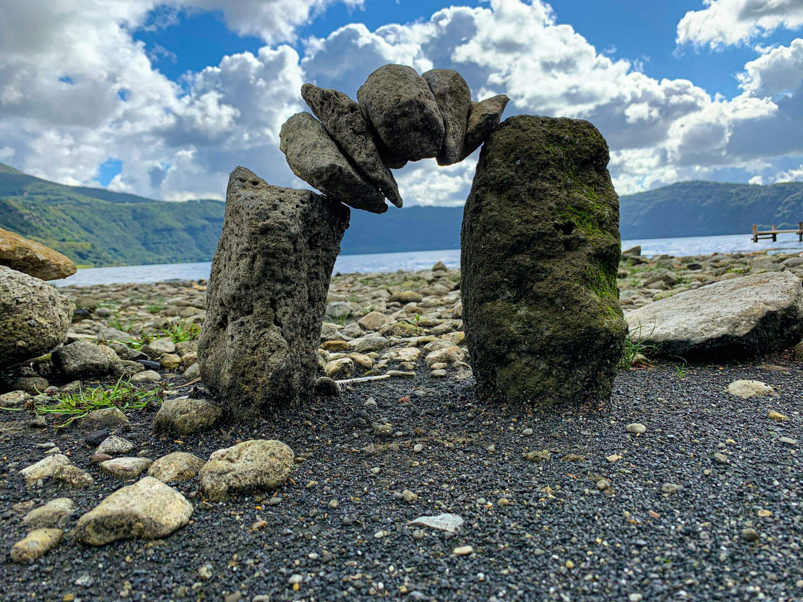 Two tall rocks with flat stones balanced on top form a mindful arch by a lake, evoking equanimity under the cloudy sky.