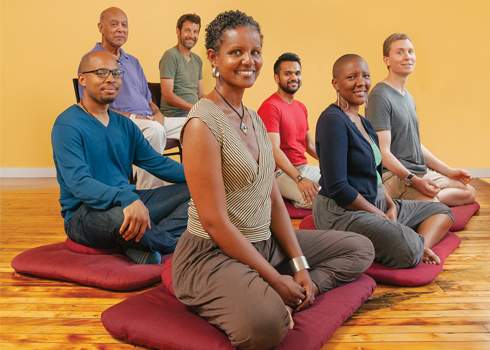 A group of seven people sits on meditation cushions arranged in two rows, smiling and relaxed. A meditation instructor guides them, with a plain yellow wall and wooden floor in the background. The individuals are diverse in appearance and dressed in comfortable, casual clothing.