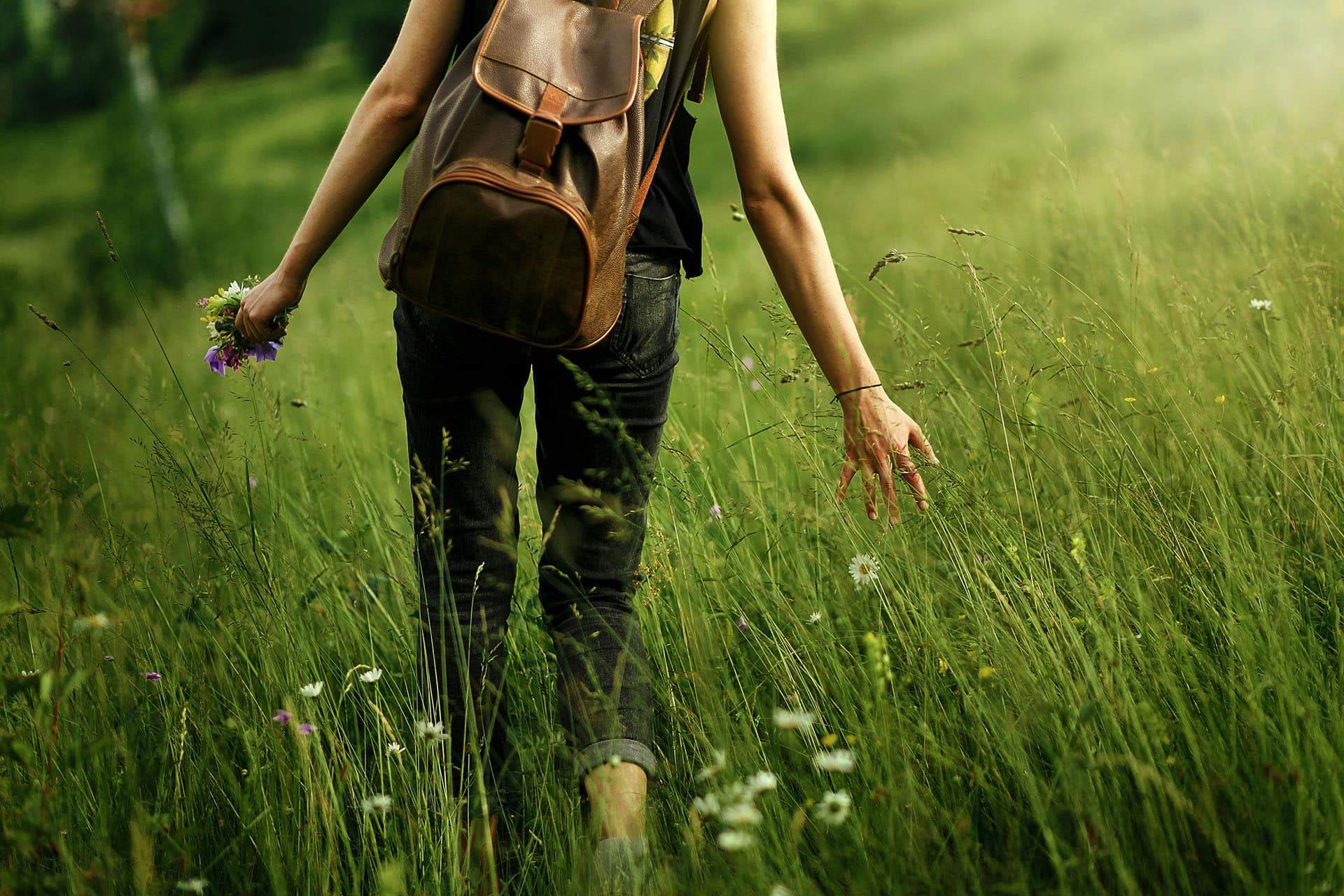 Woman practicing mindful walking among grass in meadow and holding in hand gathering wildflowers in mountains in sunlight
