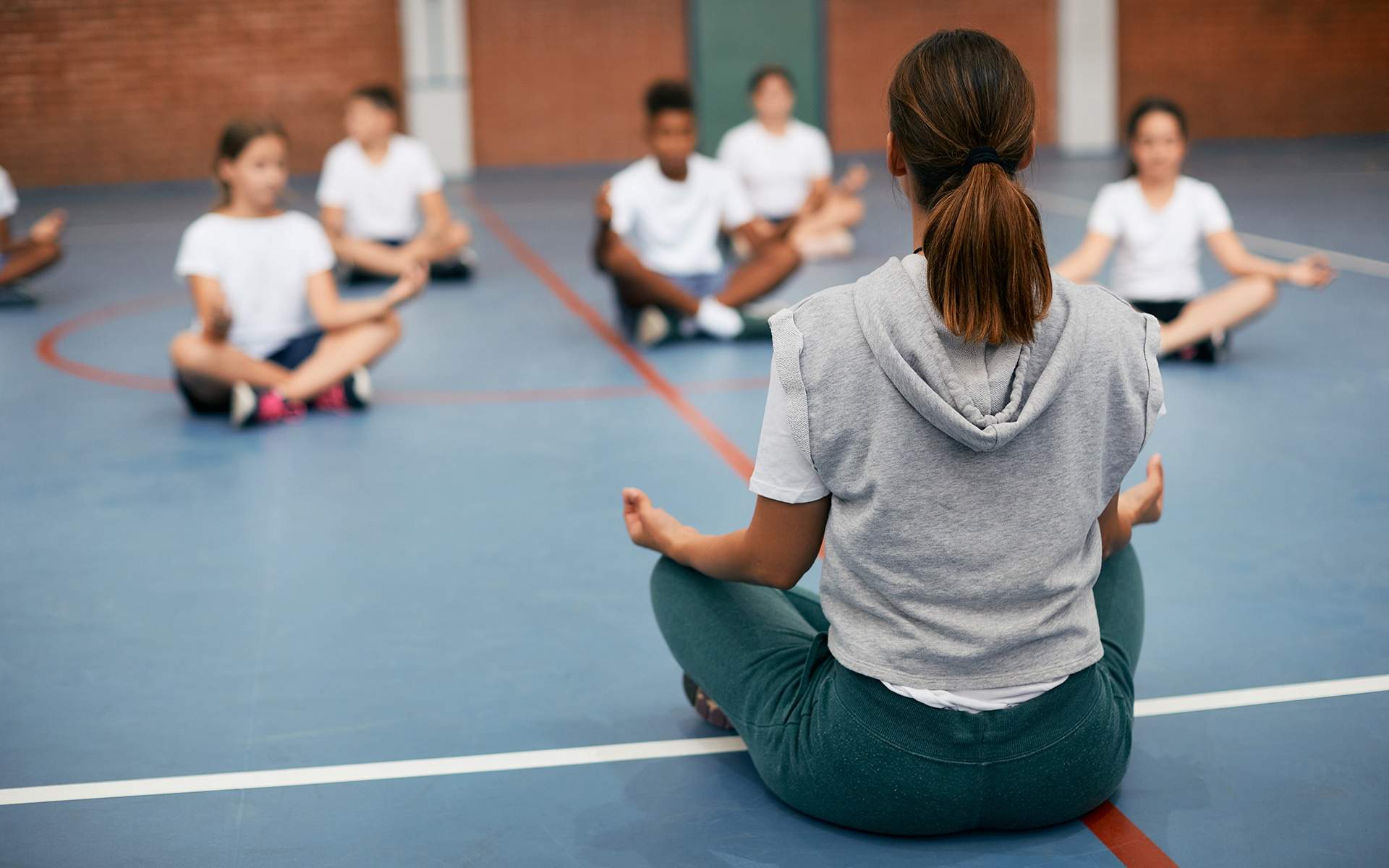 Rear view of sports teacher practicing Yoga with her students at school gym.
