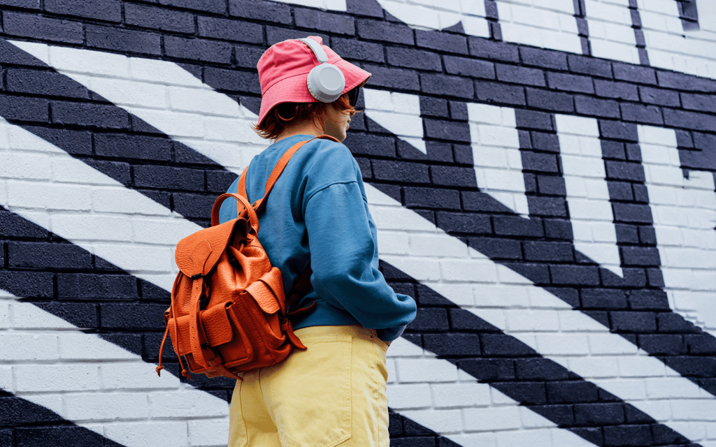 A young person wearing colorful clothes, including a bucket hat, headphones, and backpack stands in front of a painted graphic white and black brick wall. 