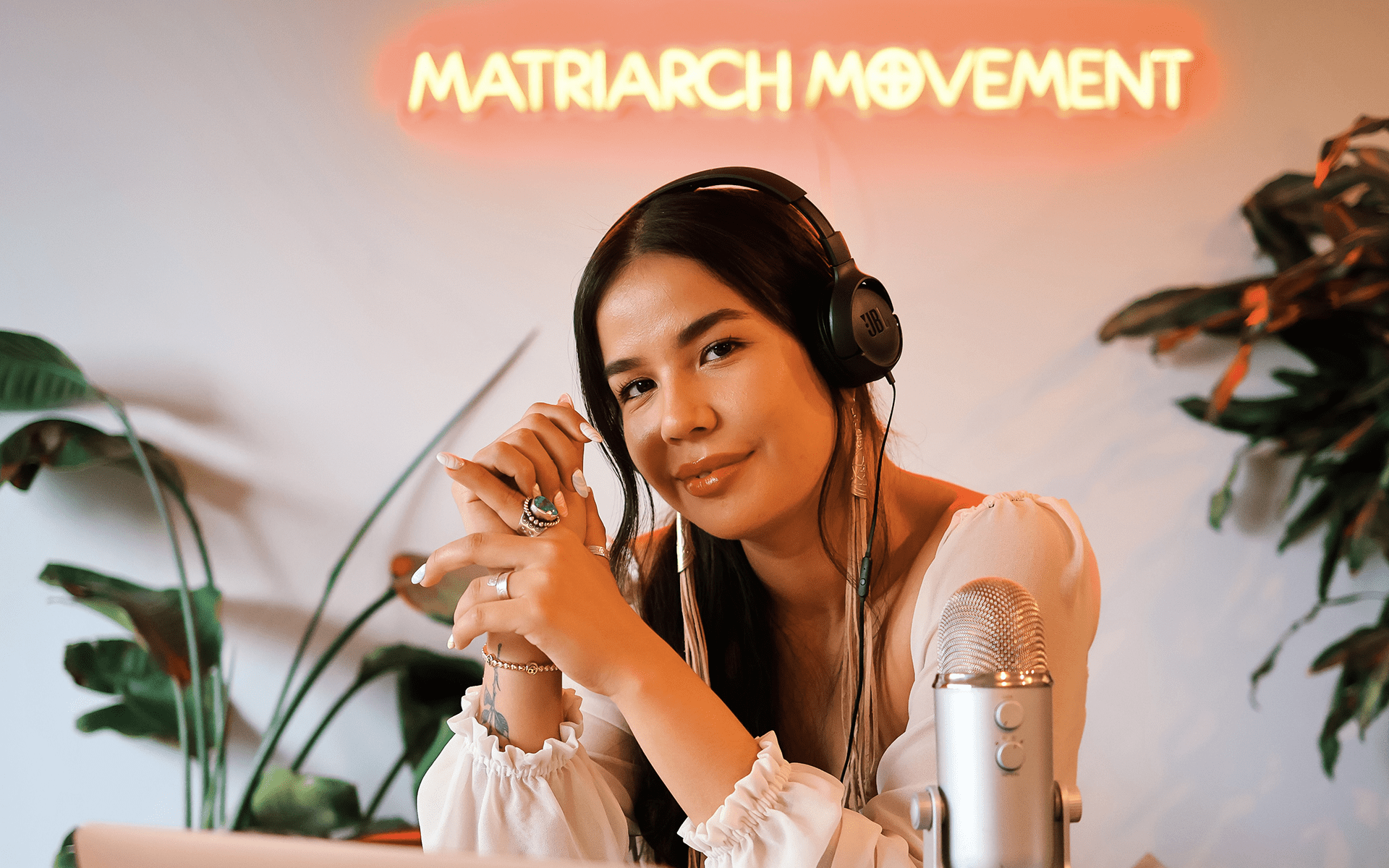 Photo of Shayla Oulette Stonechild sitting at a desk with a laptop and podcast mic with a neon sign that reads 