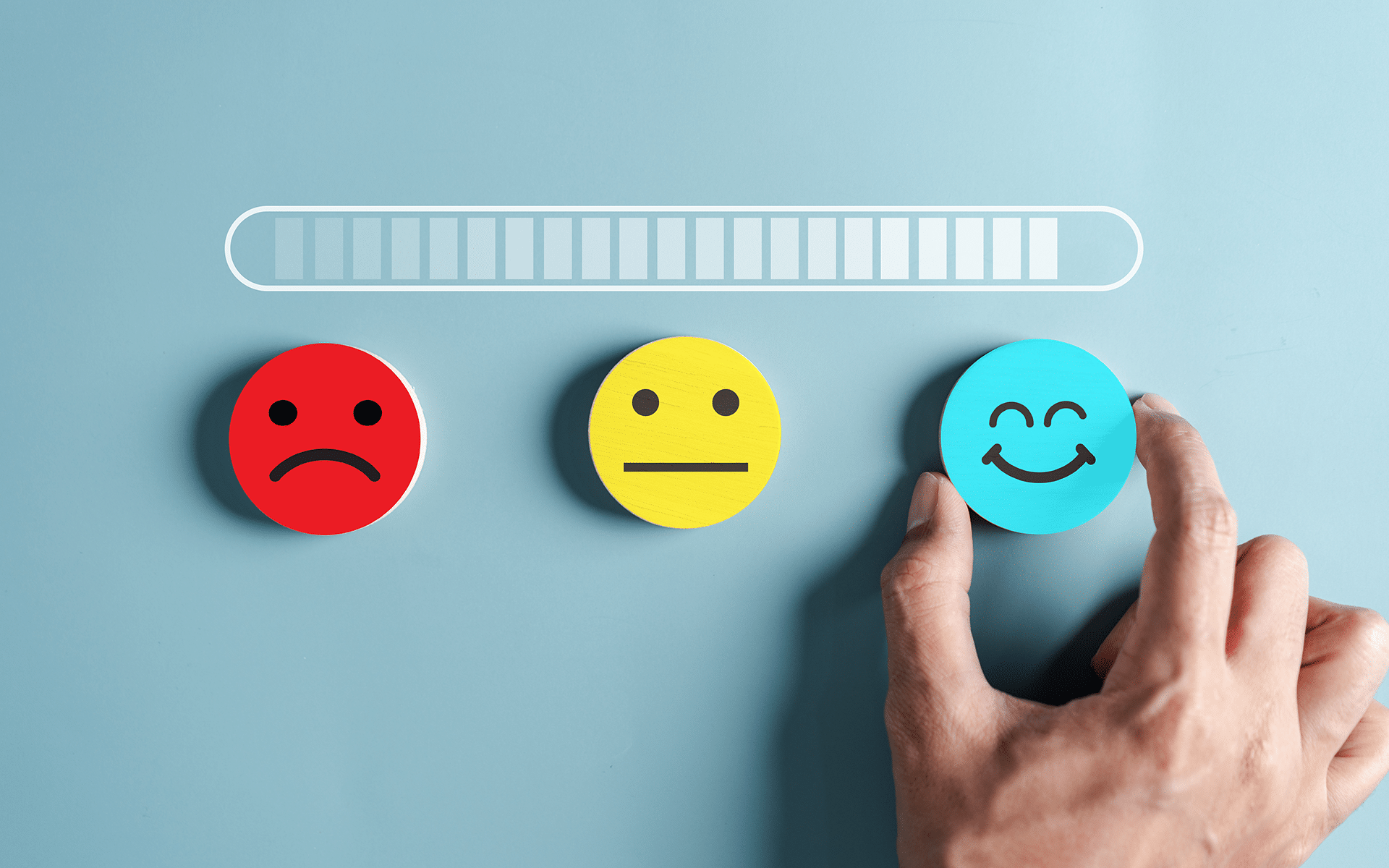 Photo of three wooden circular blocks with a variety of emotions portrayed on them sit in a row on a blue paper background. A man's hand reaches for the blue block with a smiley face.