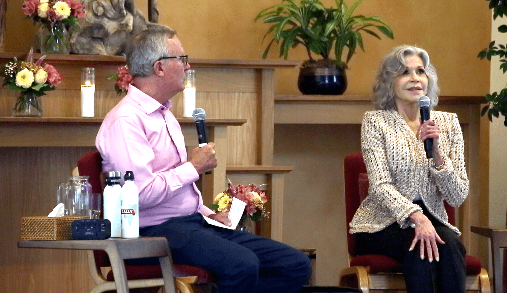 Jane Fonda sits on stage with microphones, engaged in a mindful conversation, surrounded by flowers and plants in the background.
