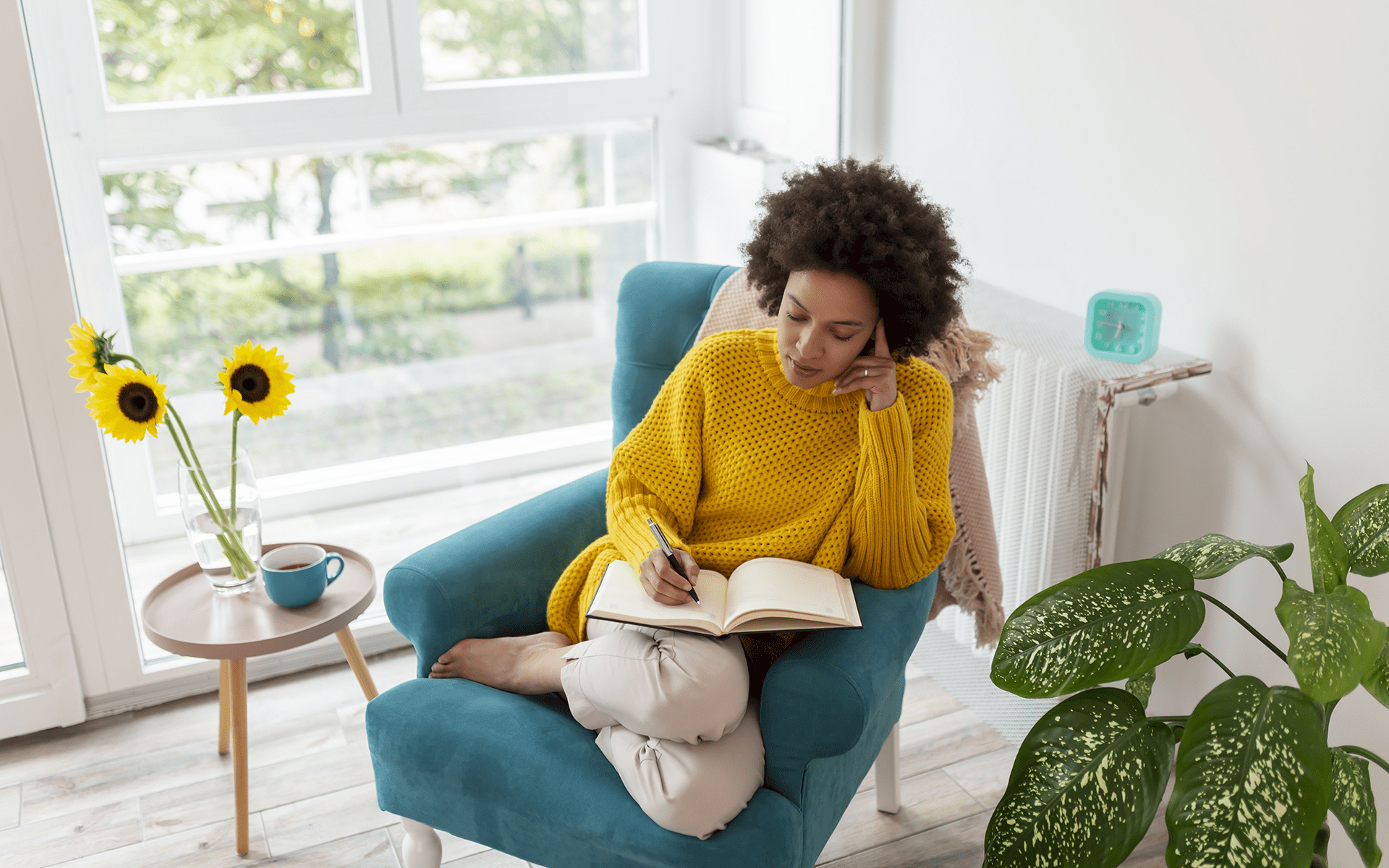 Photo of a Black woman wearing a yellow sweater and sitting in a turquoise chair writing in a journal. She's in a bright living room with sunflowers in a vase beside her.